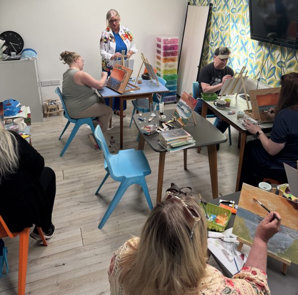 Jacqui Wheeler (stood) talking to five members of the Creative Art group who are seated painting at an easel