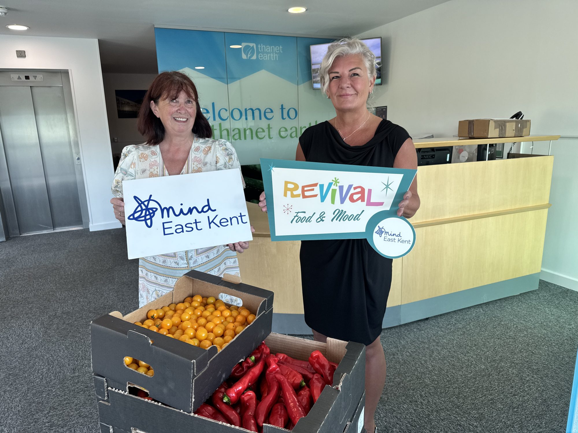 Lynne Hallowell (East Kent Mind) and Amanda Miles from Thanet Earth holding the East Kent Mind and Revival Food and Mood logos, surrounded by boxes of fresh produce including tomatoes and peppers