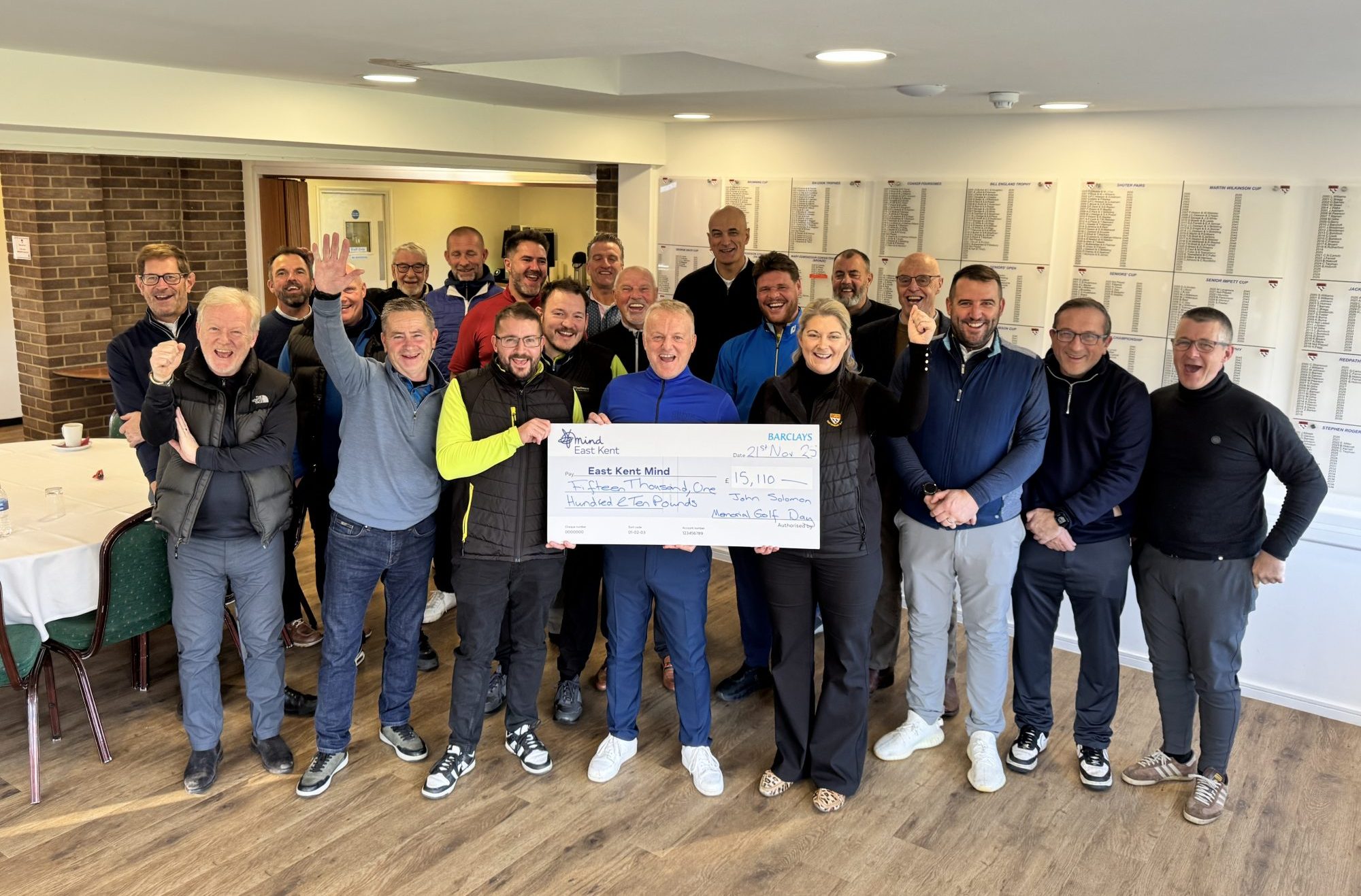 A large group of people gathered indoors, smiling and cheering as they hold a large presentation cheque made out to East Kent Mind. The group stands closely together in a golf club