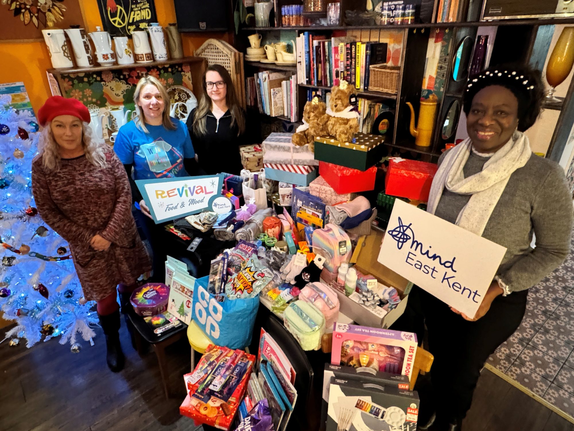 A group of people standing indoors at Revival Food & Mood in Whitstable beside a large display of donated Christmas items, including boxes, bags, toys, toiletries and food. One person holds a East Kent Mind sign and another holds a Revival Food & Mood sign, showing community partners coming together to support local people at Christmas.