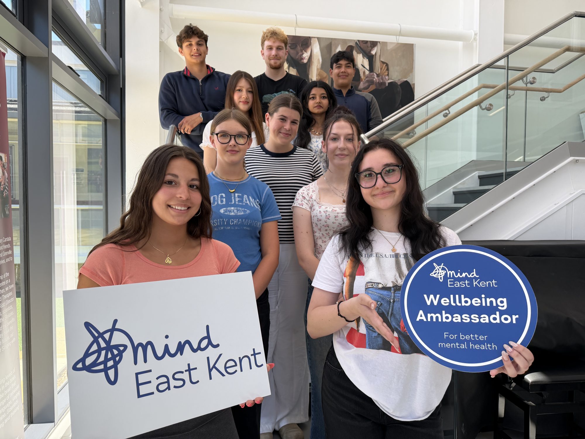 Students from Langton's are stood in a group on a staircase. Two at the front hold an East Kent Mind logo board, and a Wellbeing Ambassador banner